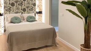Un dormitorio con cama, almohadas verdes, una planta grande en maceta y una pared con estampado floral.