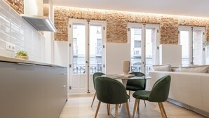 Una cocina moderna con armarios blancos, una mesa de comedor con sillas verdes y un sofá blanco en un luminoso espacio de sala de estar con pared de ladrillo y ventanas amplias.