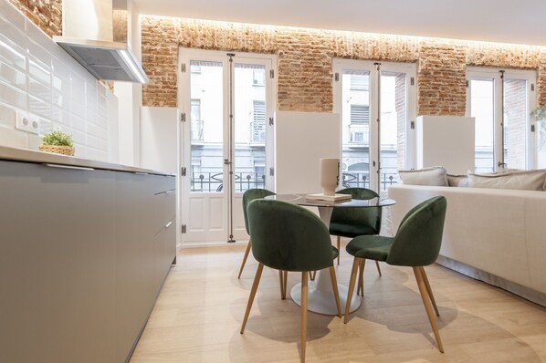 Una cocina moderna con armarios blancos, una mesa de comedor con sillas verdes y un sofá blanco en un luminoso espacio de sala de estar con pared de ladrillo y ventanas amplias.