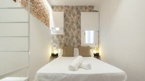 Ein ordentlich bezogenes Bett mit weißen Laken und einem zusammengerollten Handtuch, eine Wand mit gemustertem Tapetenbelag und zwei Nachttischlampen.