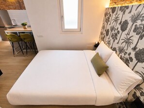 Cama com roupa de cama branca, um travesseiro verde e uma mesa de cabeceira com abajur. Há uma janela sobre a cama e uma parede com padrão de palmeiras.