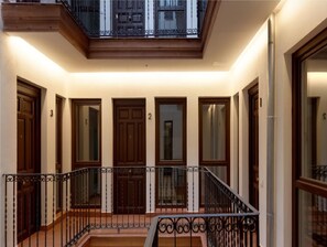 Un pasillo con puertas de madera numeradas del 2 al 5, un balcón con barandilla de hierro forjado y una escalera que sube a un piso superior.