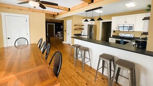 Een moderne keuken met houten balken, een eethoek met een lange tafel en stoelen, en een bar met krukjes.