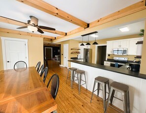 A modern kitchen with wooden beams, a dining area with a long table and chairs, and a bar area with stools.