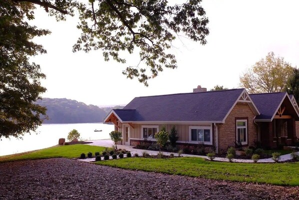 A two-story house with a stone facade, a dark roof, and a front porch. It is situated by a body of water with a view of hills in the background.