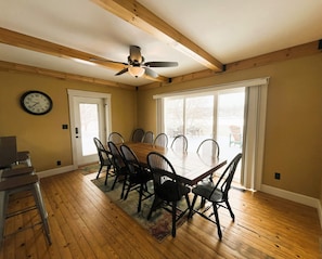 A dining room with a wooden table and chairs, a ceiling fan, a clock, and large windows.