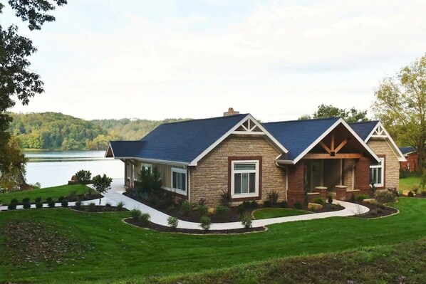 A two-story house with a stone facade and a blue roof, situated by a lake with a well-maintained lawn and a curved driveway.