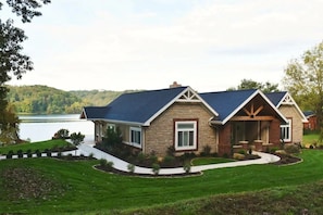 A two-story house with a stone facade and blue roof, surrounded by a well-maintained lawn and a lake view. - Lake House Villa by HHSW (Logan)
