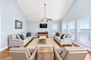 A living room with a high ceiling, a ceiling fan, a flat-screen TV, and a large window with blinds.