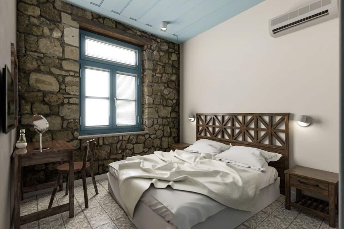 A bedroom with a stone wall, a wooden headboard, a bed with white bedding, a small table with a mirror, and a window with blue shutters.