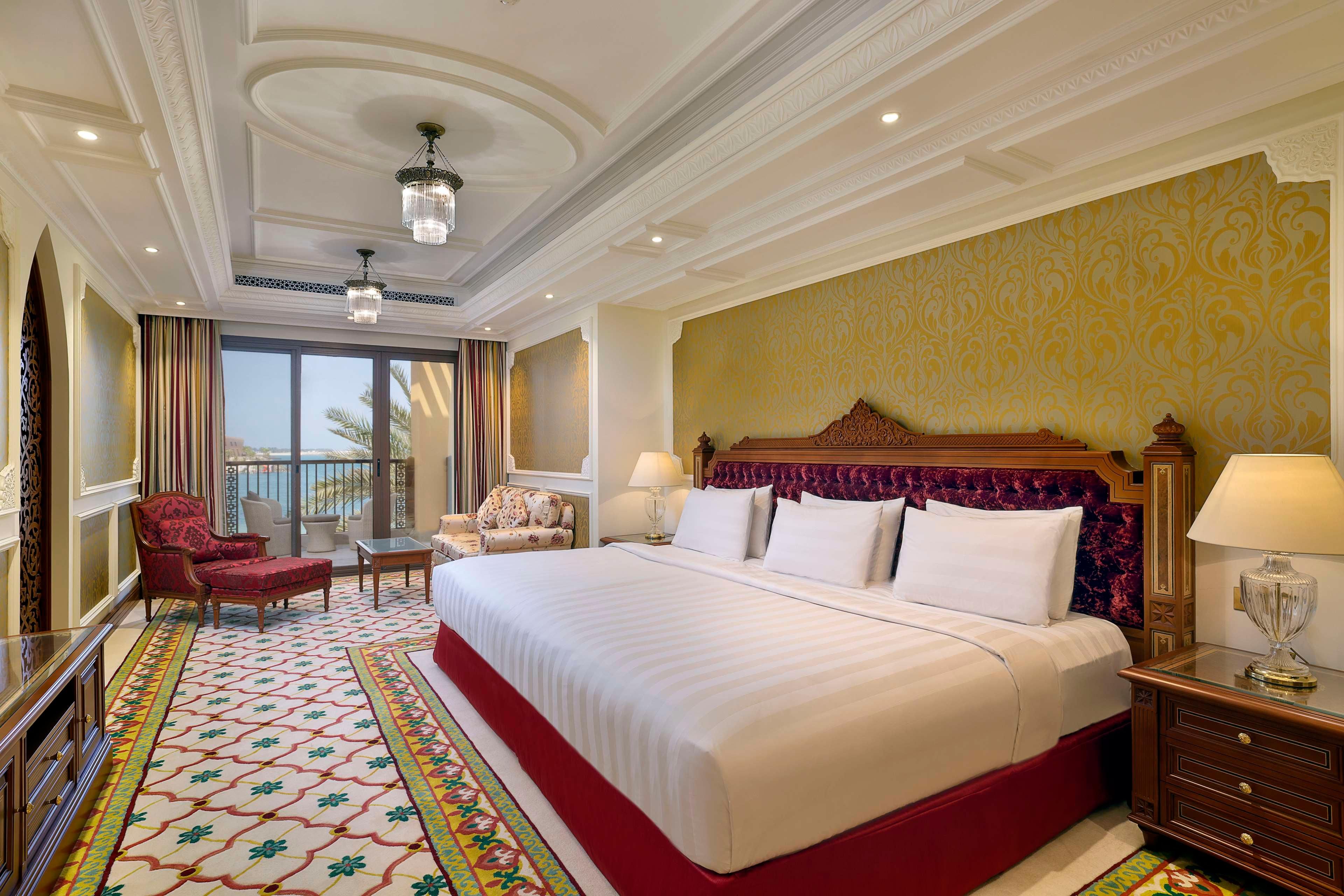 A spacious hotel room with a large bed, a sitting area with a chair and a small table, a balcony with a view, and a decorative carpet.