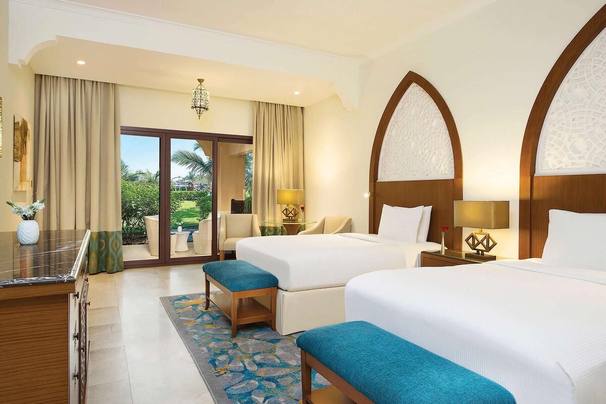 A hotel room with two beds, a desk, a chair, and a view of the outdoors.