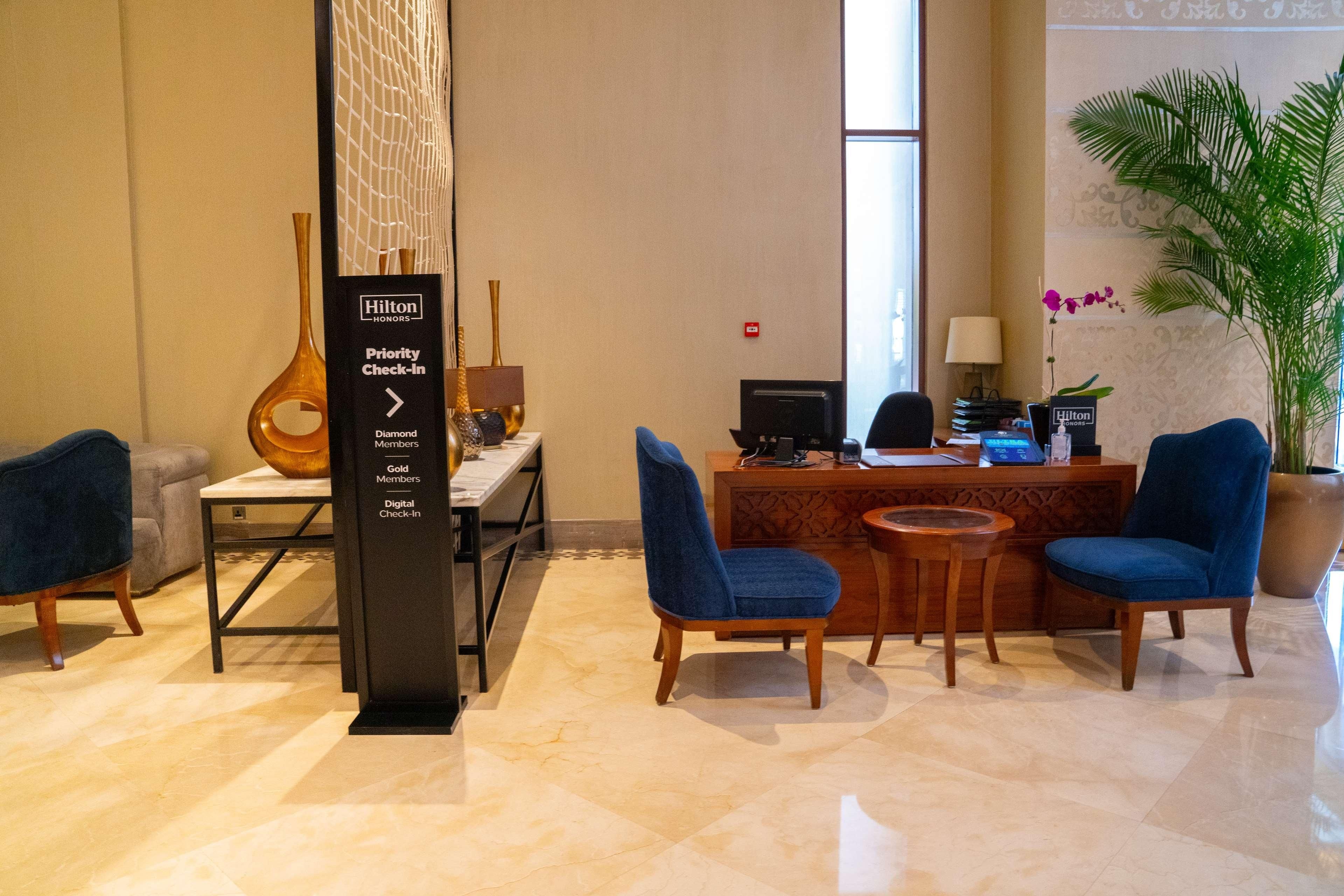 A hotel reception area with a priority check-in sign, a reception desk, two blue chairs, a wooden table, and a potted plant.