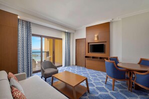 Ein Hotelzimmer mit einer Couch, einem kleinen Tisch, einem an einem Holzkasten montierten Fernseher, einem Balkon mit Blick und einem gemusterten Teppich.