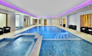 A large indoor swimming pool with a clear blue water surface, surrounded by black lounge chairs and a marble wall with a decorative panel.