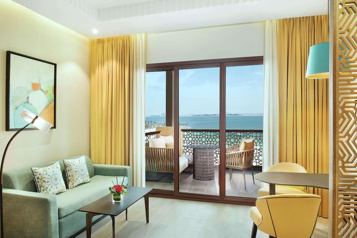 Een hotelkamer met een bank, een tafel, een stoel, een balkon met uitzicht op zee en een groot raam.