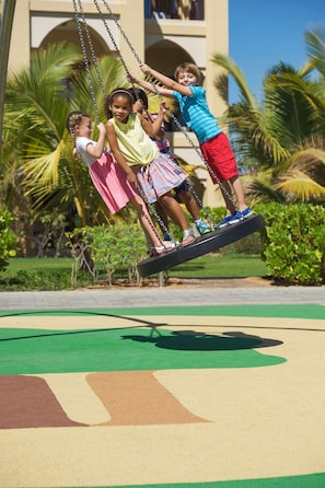 Children on a swing set in a park with palm trees and a building in the background. - Doubletree by Hilton Resort & Spa Marjan Island (Ras Al Khaimah)