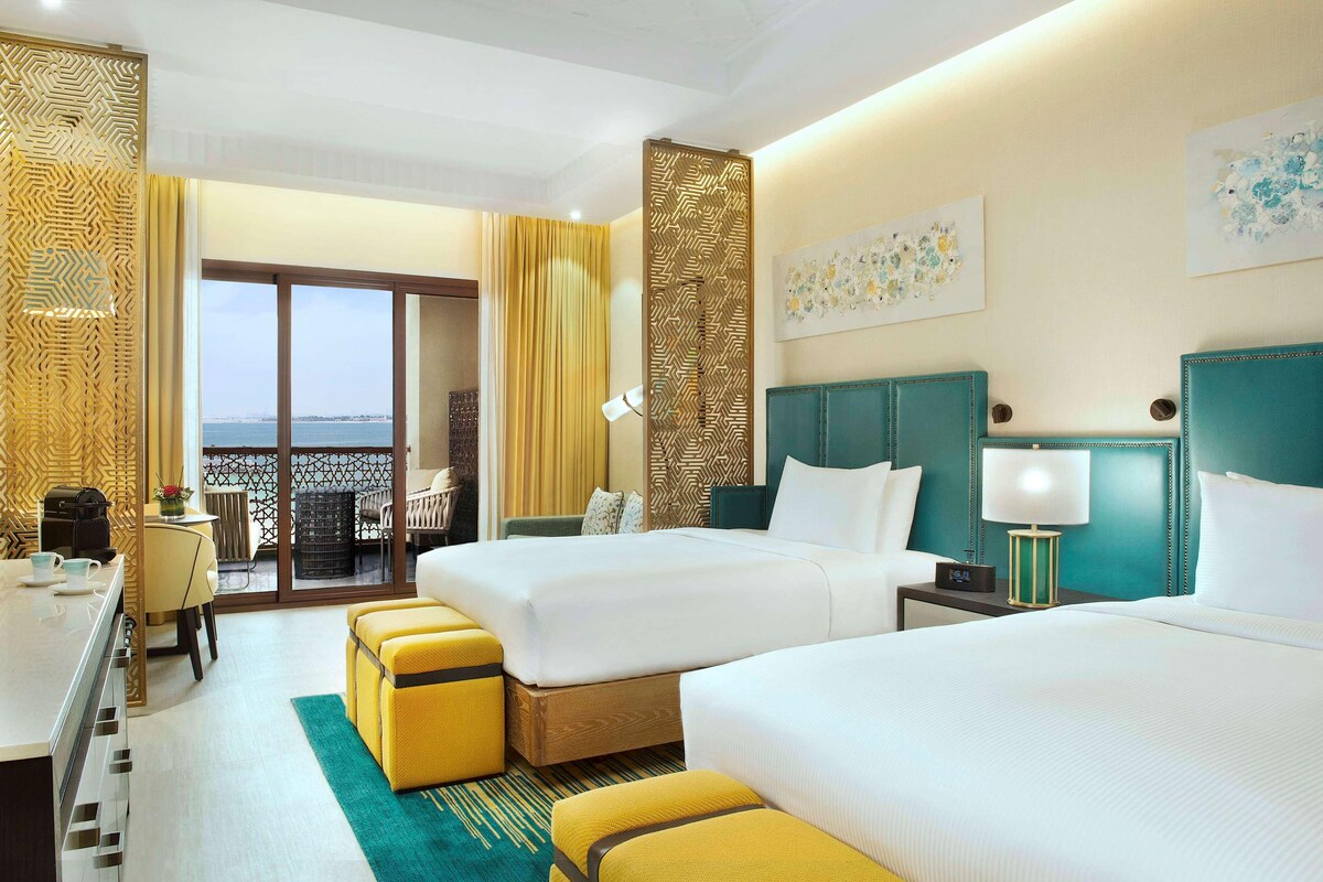 A hotel room with two beds, a balcony with a view, a desk with a chair, and a dining area.