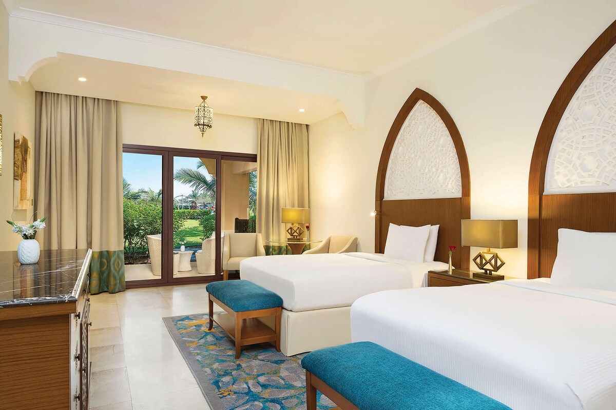 A hotel room with two beds, a desk, and a view of the outdoors.