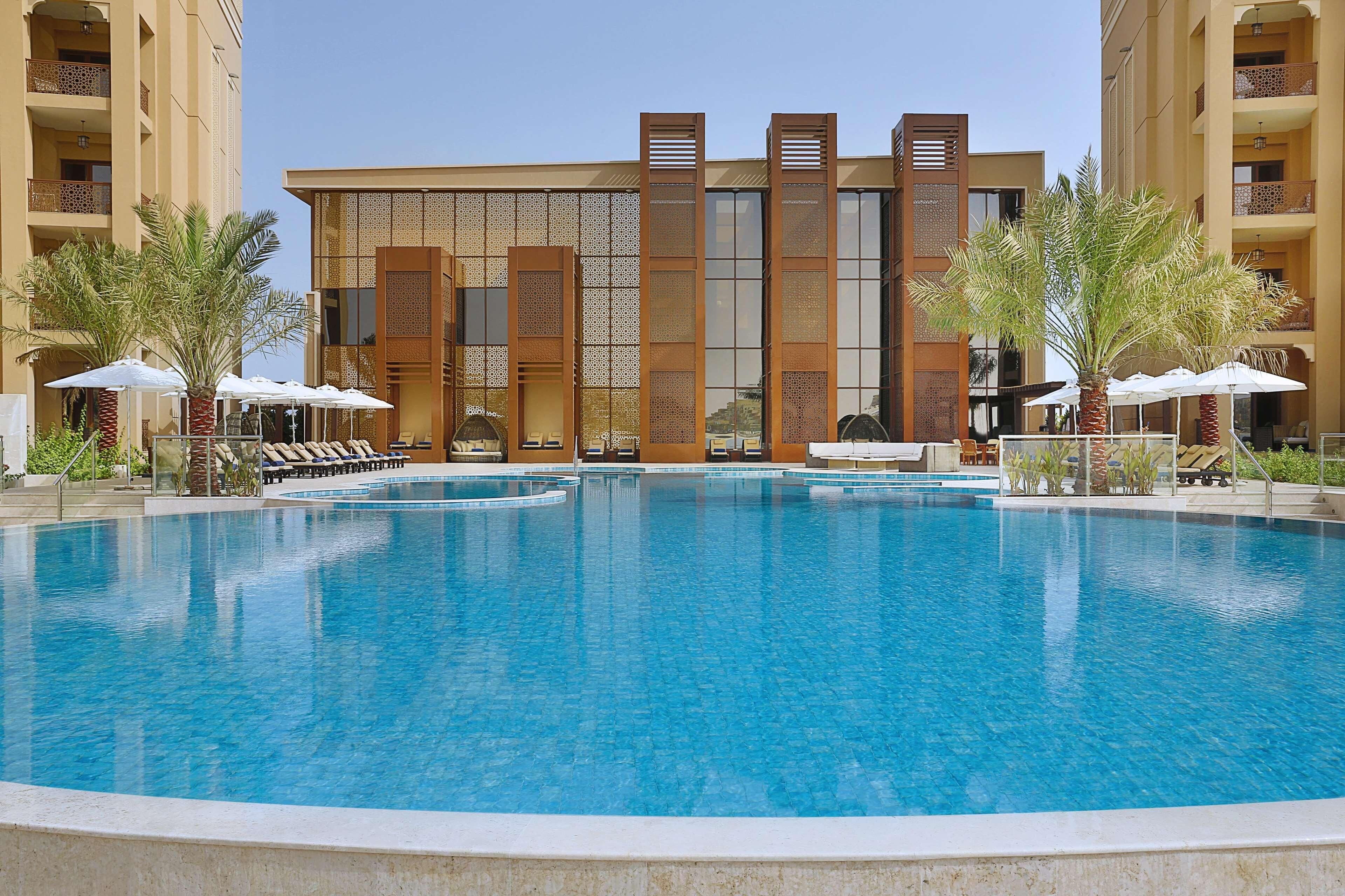 A hotel with a large pool, sun loungers, and palm trees.