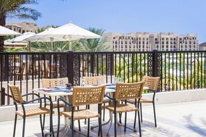 Un área de comedor al aire libre con sillas y mesas de mimbre, sombrillas y una vista de un edificio al fondo.