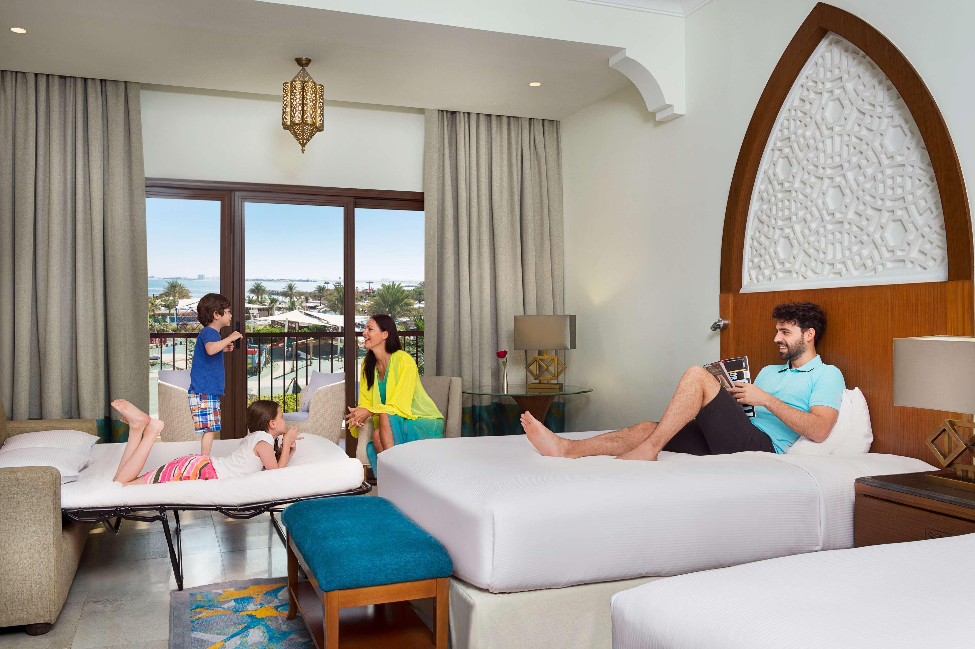 A hotel room with a large window, a bed, a massage table, and a small table with a lamp.
