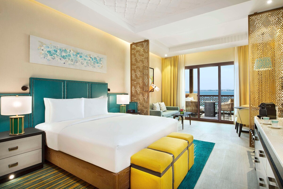 A hotel room with a large bed, a desk, a chair, a sofa, and a view of the sea through the window.