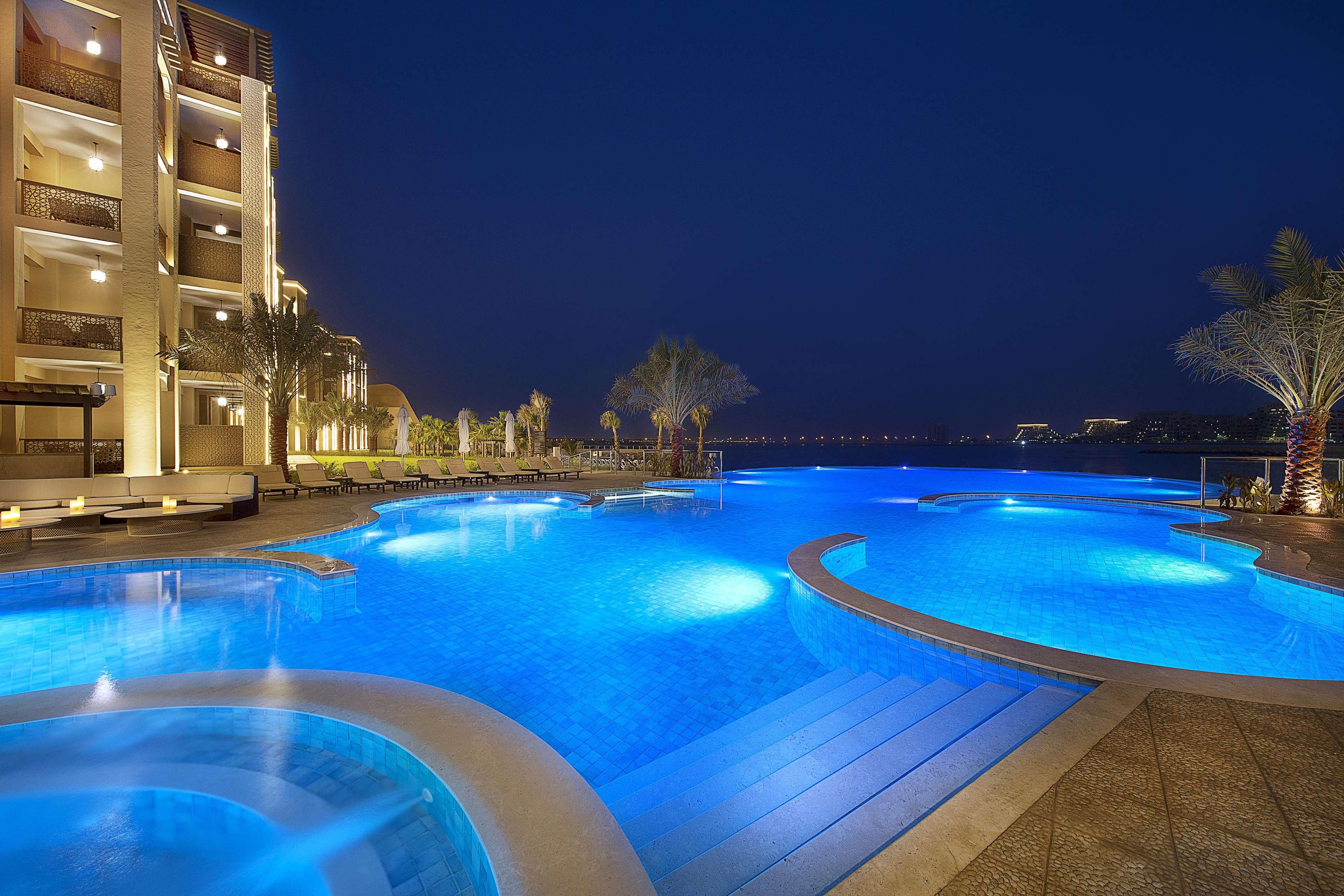 A nighttime swimming pool area with multiple levels of a building adjacent to it, and a view of the city skyline in the background.