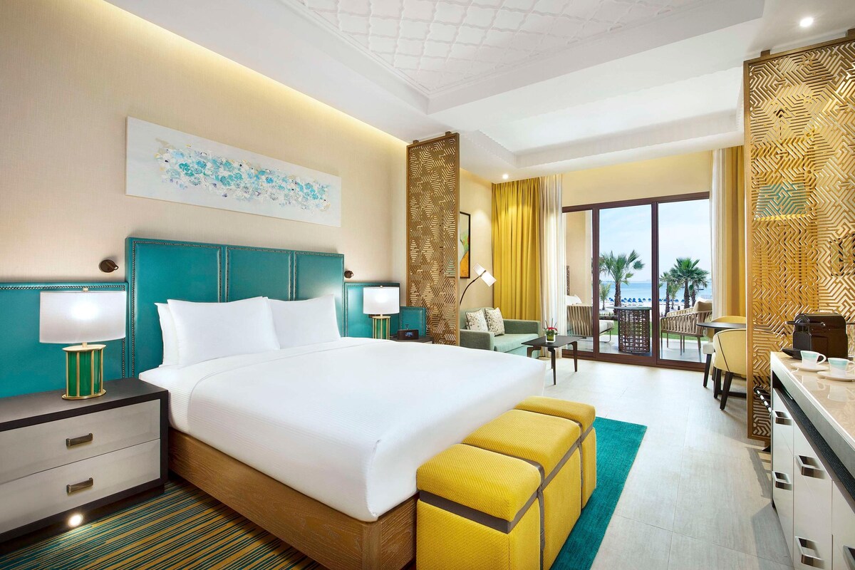 A hotel room with a bed, bedside tables, a desk, a chair, a sofa, a balcony with a view, and a decorative wall panel.