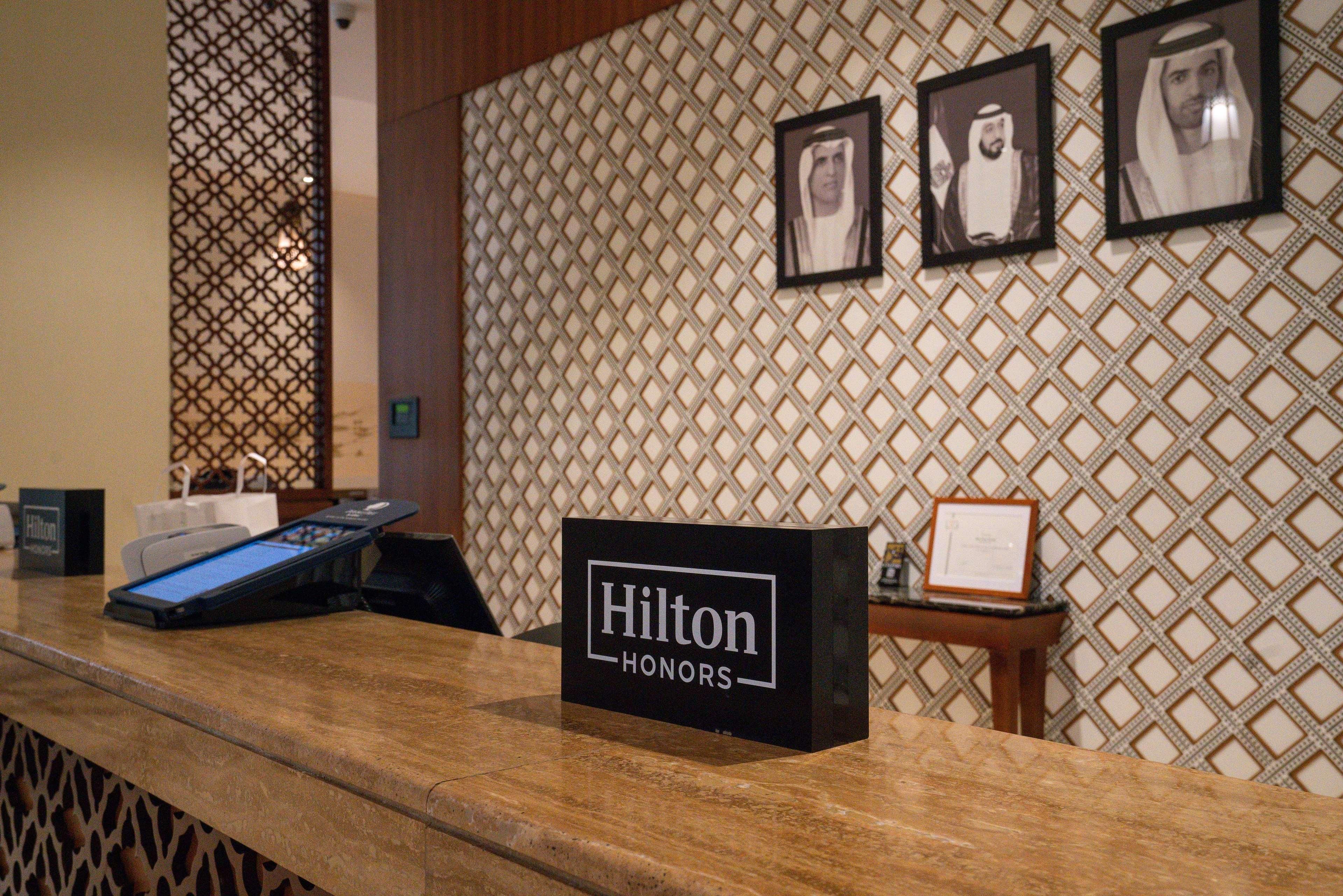 A hotel reception desk with a 'Hilton Honors' sign, a digital check-in kiosk, and framed photographs on the wall.