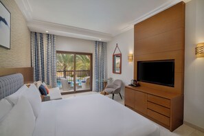 A hotel room with a large bed, a TV, a seating area with a table and chairs, and a balcony with a pool view.