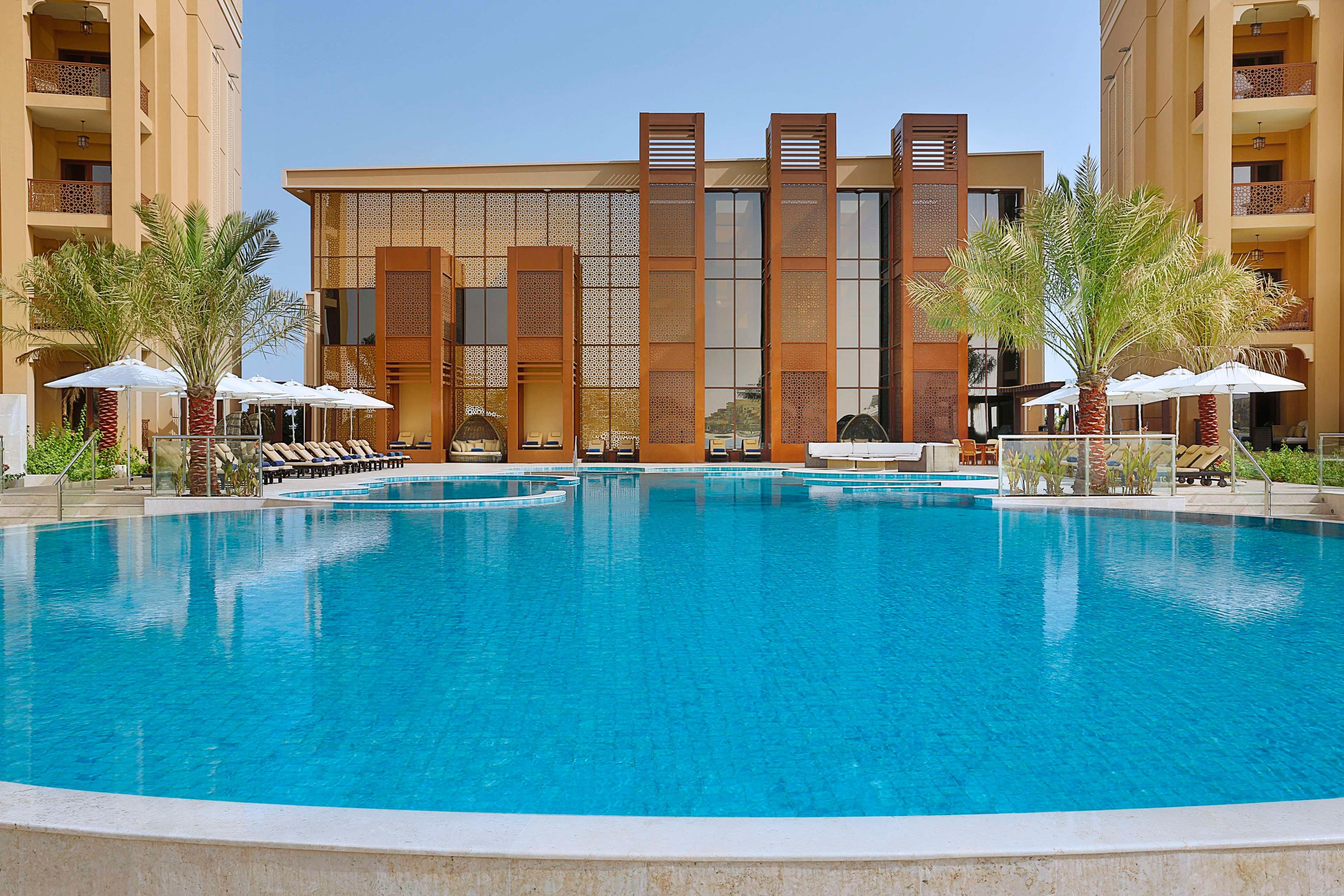 A hotel with a large swimming pool, sun loungers, and palm trees.