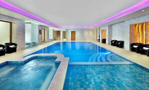 A large indoor swimming pool with a tiled floor, surrounded by black lounge chairs and a marble wall with a decorative panel. - Doubletree by Hilton Resort & Spa Marjan Island (Ras Al Khaimah)