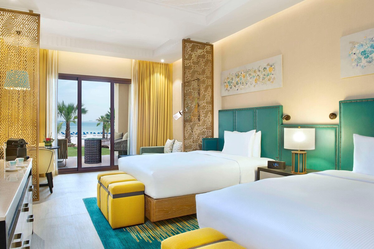A hotel room with two beds, a desk, a chair, and a view of the beach.