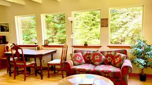 A cozy living room with a wooden dining table, chairs, a patterned sofa, and a small round table with papers. Large windows offer views of greenery outside. - 50+ acre Nature Preserve with Ponds & Meadows: Azul Cottage Retreat (Mendocino)