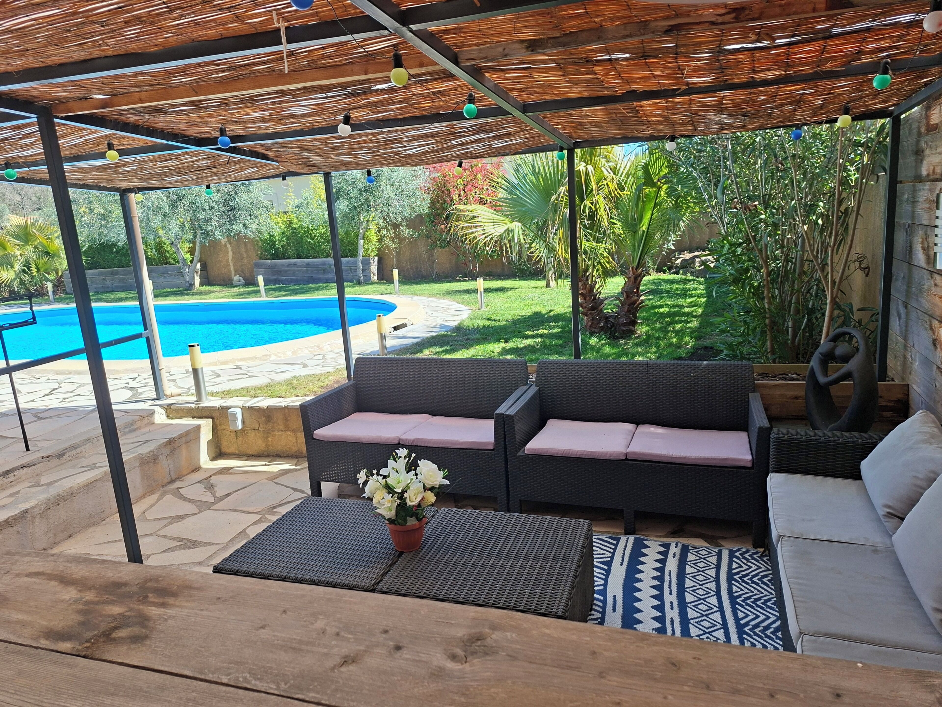 Un espacio de estar techado al aire libre con mobiliario de mimbre, piscina y abundante vegetación.