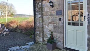 A stone building with a white door, a small garden area with a chair and a potted plant, and a sign reading "Hetton Byre".