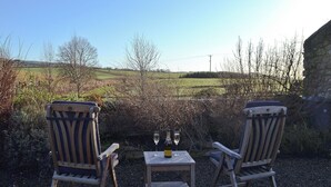 Two wooden chairs facing a table with a bottle and a glass, set against a rural landscape with fields and trees.