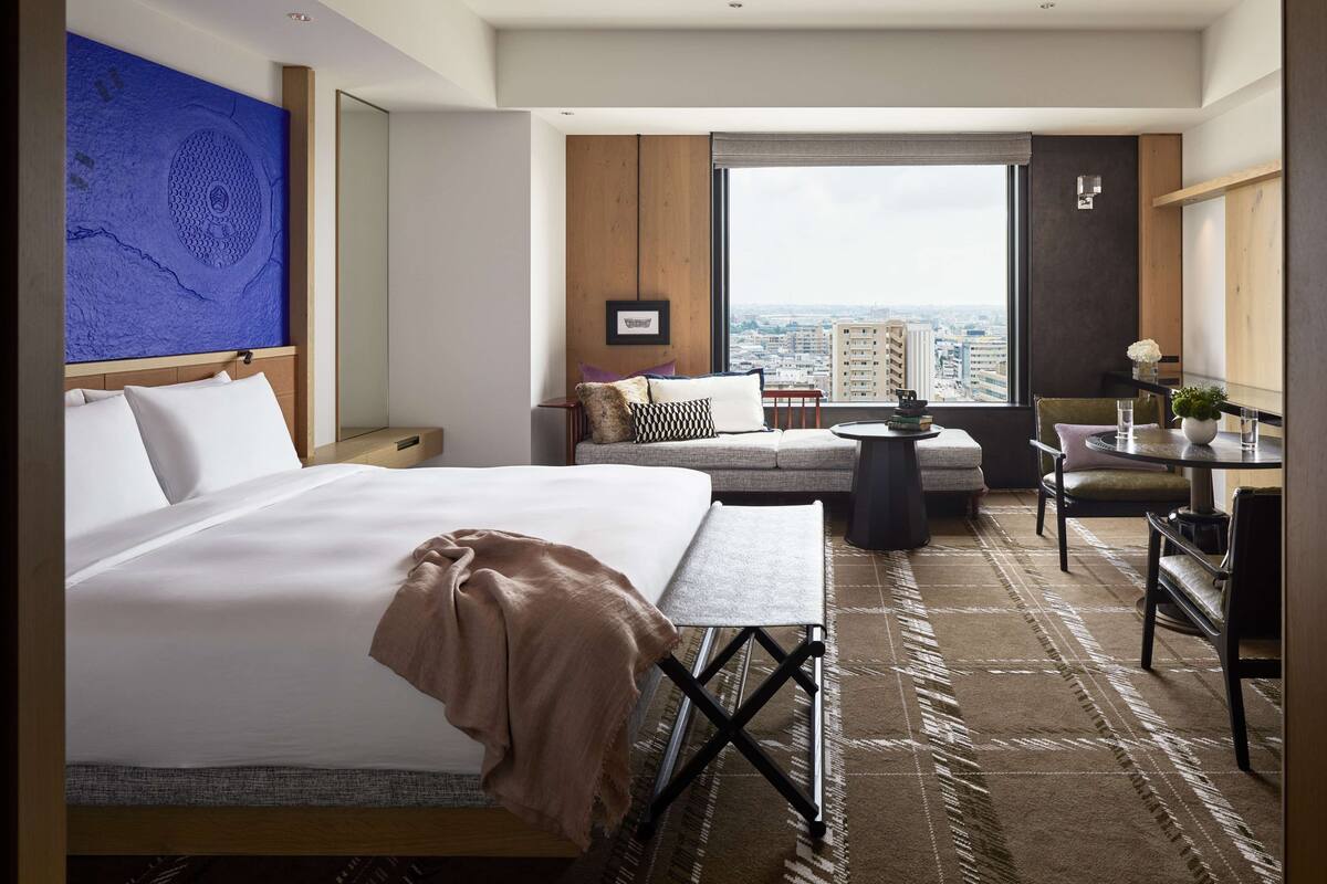 Ein Hotelzimmer mit einem großen Bett, einem kleinen Tisch, einer Sitzecke und Blick auf die Stadt.