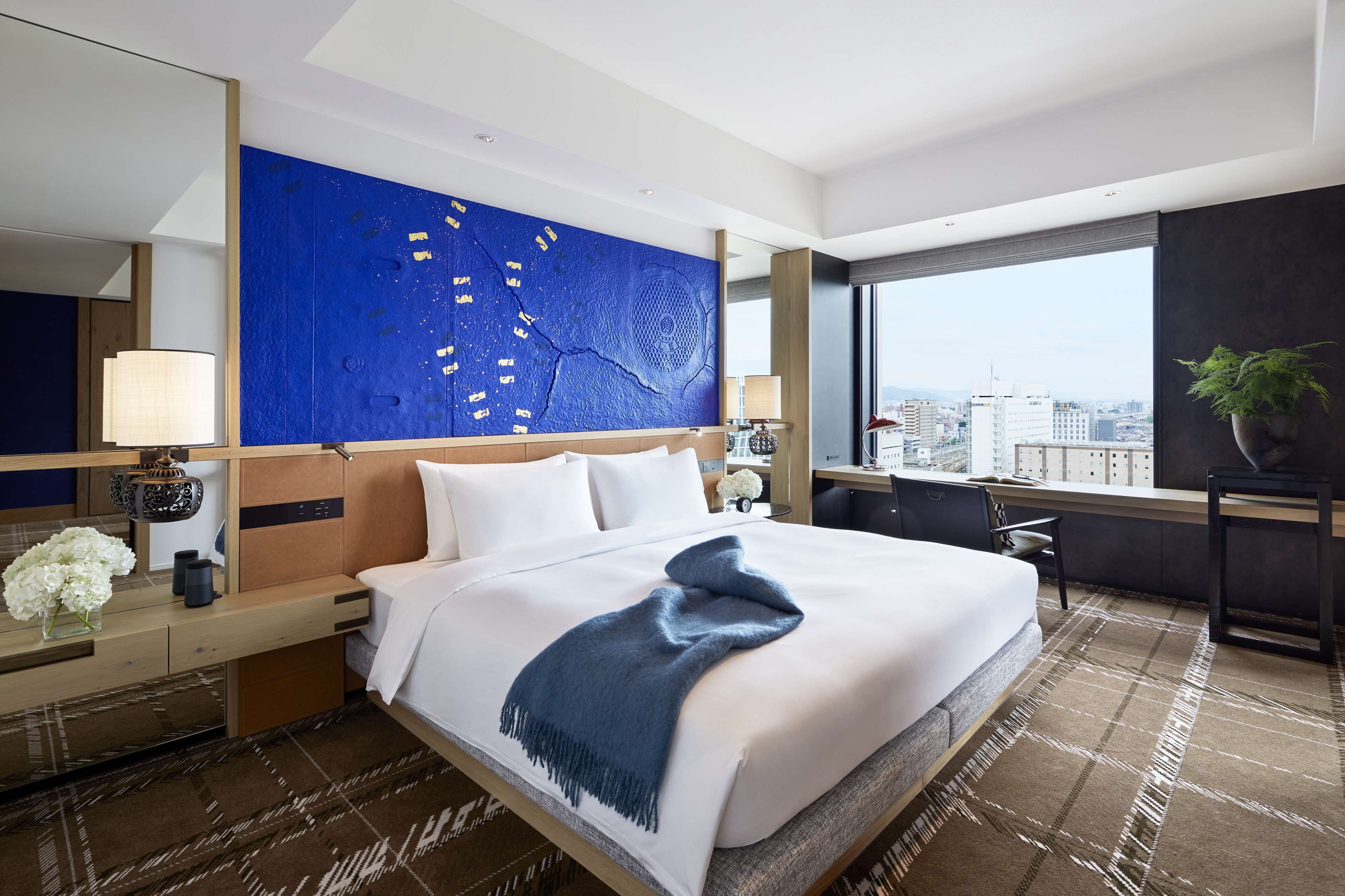 A modern hotel room with a large bed, a city view, a blue abstract wall art, and a desk with a lamp.
