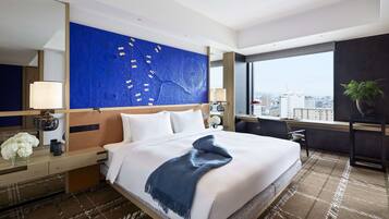 A modern hotel room with a large bed, a city view, a blue abstract wall art, and a desk with a lamp.