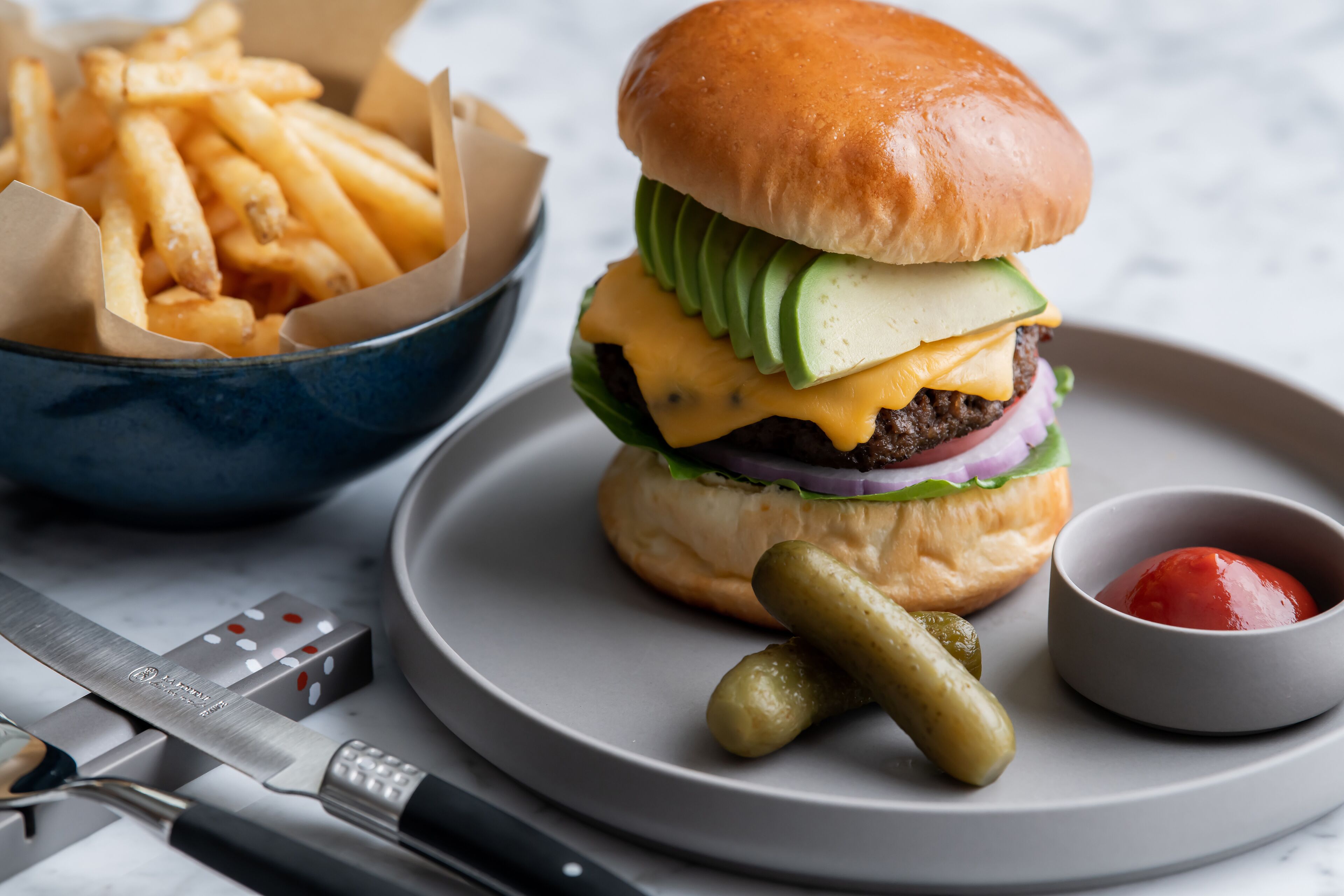 Een hamburger met augurken en ketchup op een bord, een bak friet en een mes op een tafel.