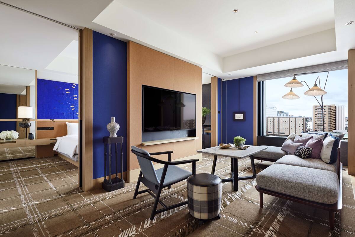 Ein modernes Hotelzimmer mit großem Fernseher, bequemen Sitzgelegenheiten, einer Essecke und Blick auf die Stadt.