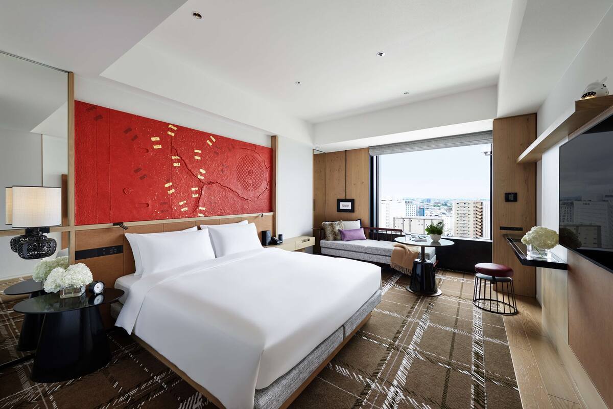Ein modernes Hotelzimmer mit einem großen Bett, einer roten Akzentwand, Blick auf die Stadt und einer kleinen Sitzecke mit Tisch und Stühlen.