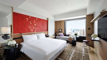 A modern hotel room with a large bed, a red accent wall, a city view, and a small seating area with a table and chairs.