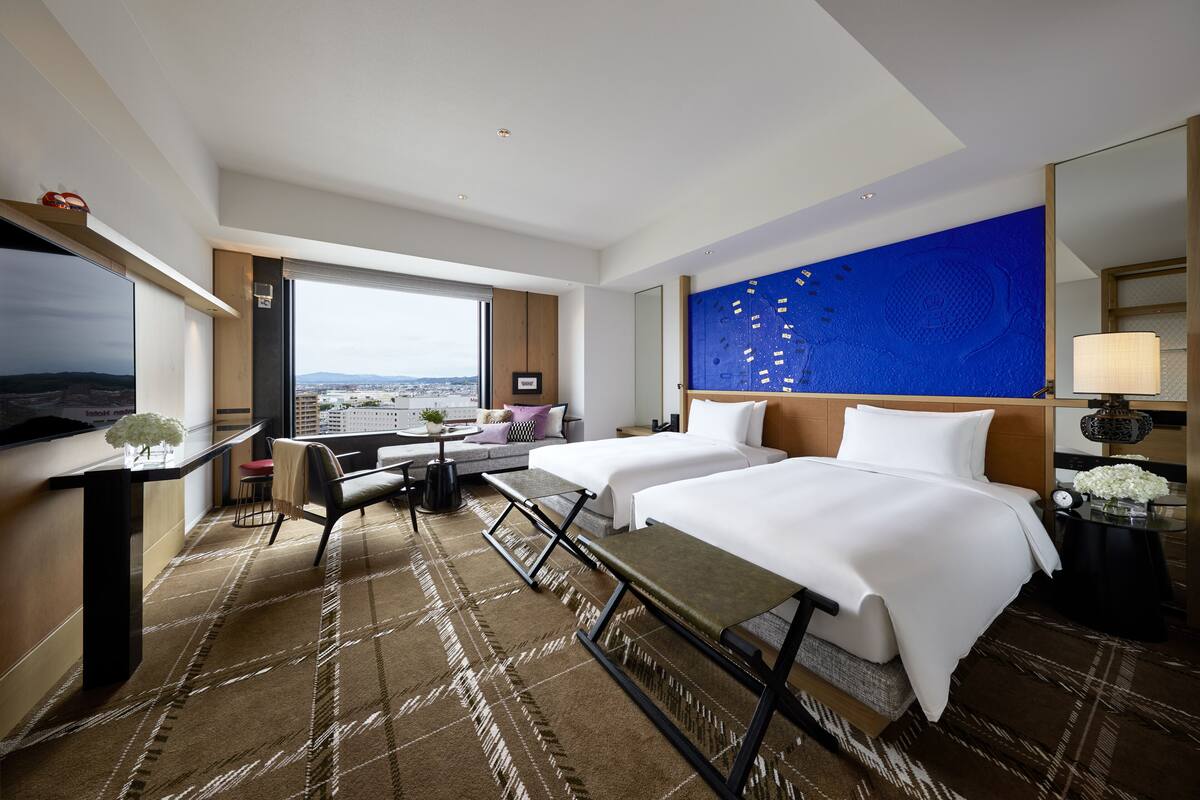 Ein Hotelzimmer mit zwei Betten, einem Schreibtisch, einem Stuhl, einem großen Fenster mit Stadtblick und einer blauen Akzentwand.