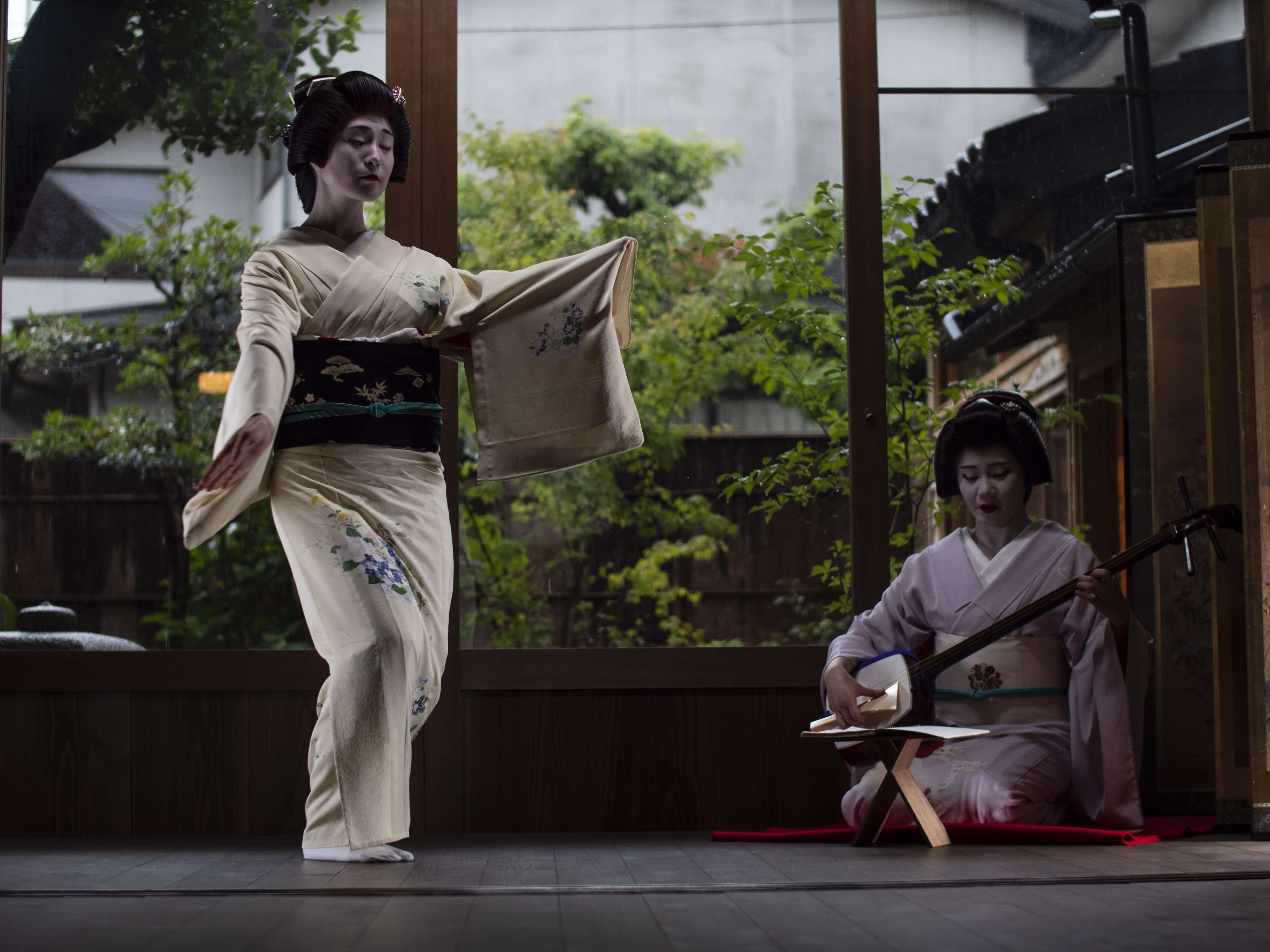 Two individuals in traditional attire, one standing and one seated, in an indoor setting with large windows and a view of greenery outside.