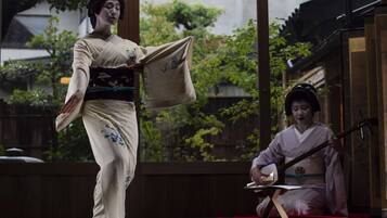 Two individuals in traditional attire, one standing and one seated, in an indoor setting with large windows and a view of greenery outside.