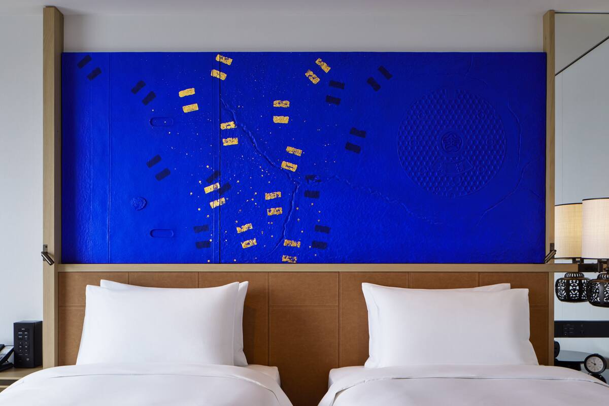 Ein Hotelzimmer mit großem, blauen Kunstwerk an der Wand, einem hölzernen Kopfteil, zwei weißen Betten und einem Nachttisch mit Lampe und Uhr.