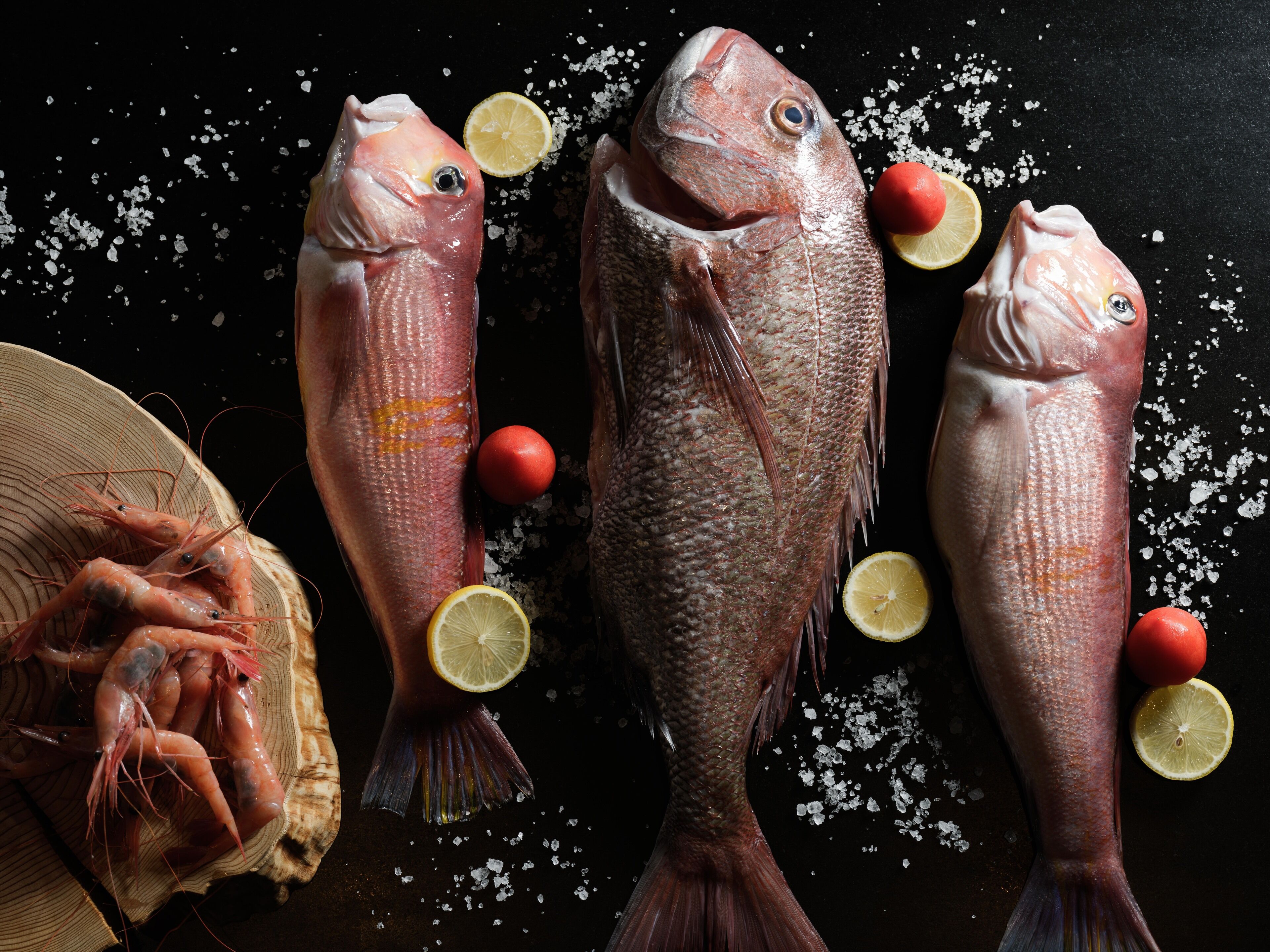 Trois poissons et quelques crevettes sur une surface sombre, parsemée de sel et de tranches de citron.